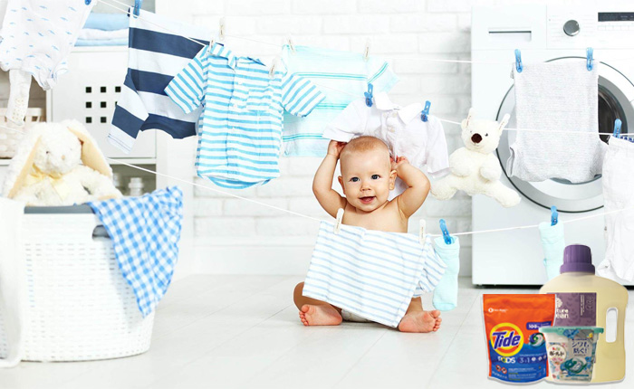 A baby in blue and white striped clothes happily raises his hands on the laundry room floor, surrounded by children's clothes hanging on the clothesline and washing liquid leaning against the washing machine